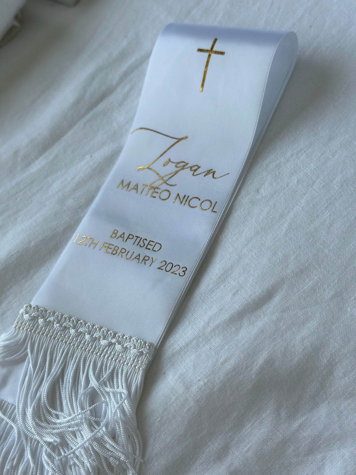 A white baptismal Stole with gold foil lettering and a gold cross, laid diagonally on a white textured fabric background. The sash features the name 'Logan', the Christian name 'Matteo Nicol', the baptism date '19th February 2023', and a decorative fringed tassel at the bottom.