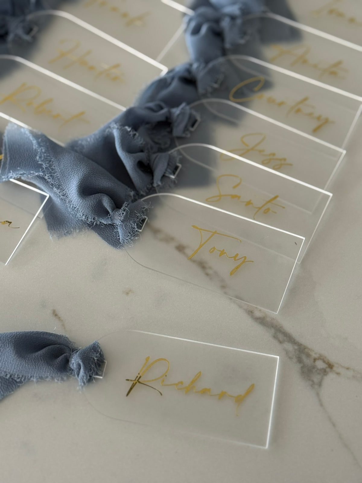 Clear acrylic place cards with gold foil script names, tied with dusty blue ribbon, arranged on a light marble surface.