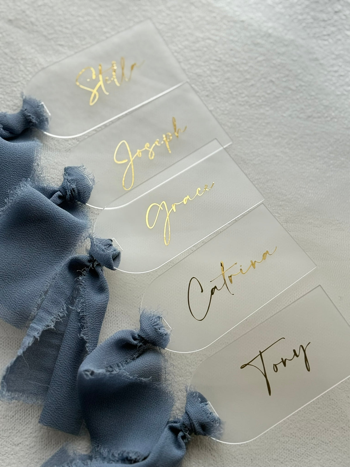 Five clear acrylic place cards with gold cursive names, attached to dusty blue ribbon ties, arranged on a textured white surface.