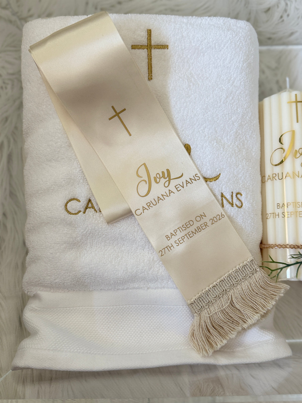 A white bath towel with gold embroidered cross and lettering, draped with a cream satin baptism sash that reads 'Joy Cariana Evans Baptised on 27th September 2025.' A matching cream candle is partially visible on the right, set on a soft white textured surface.