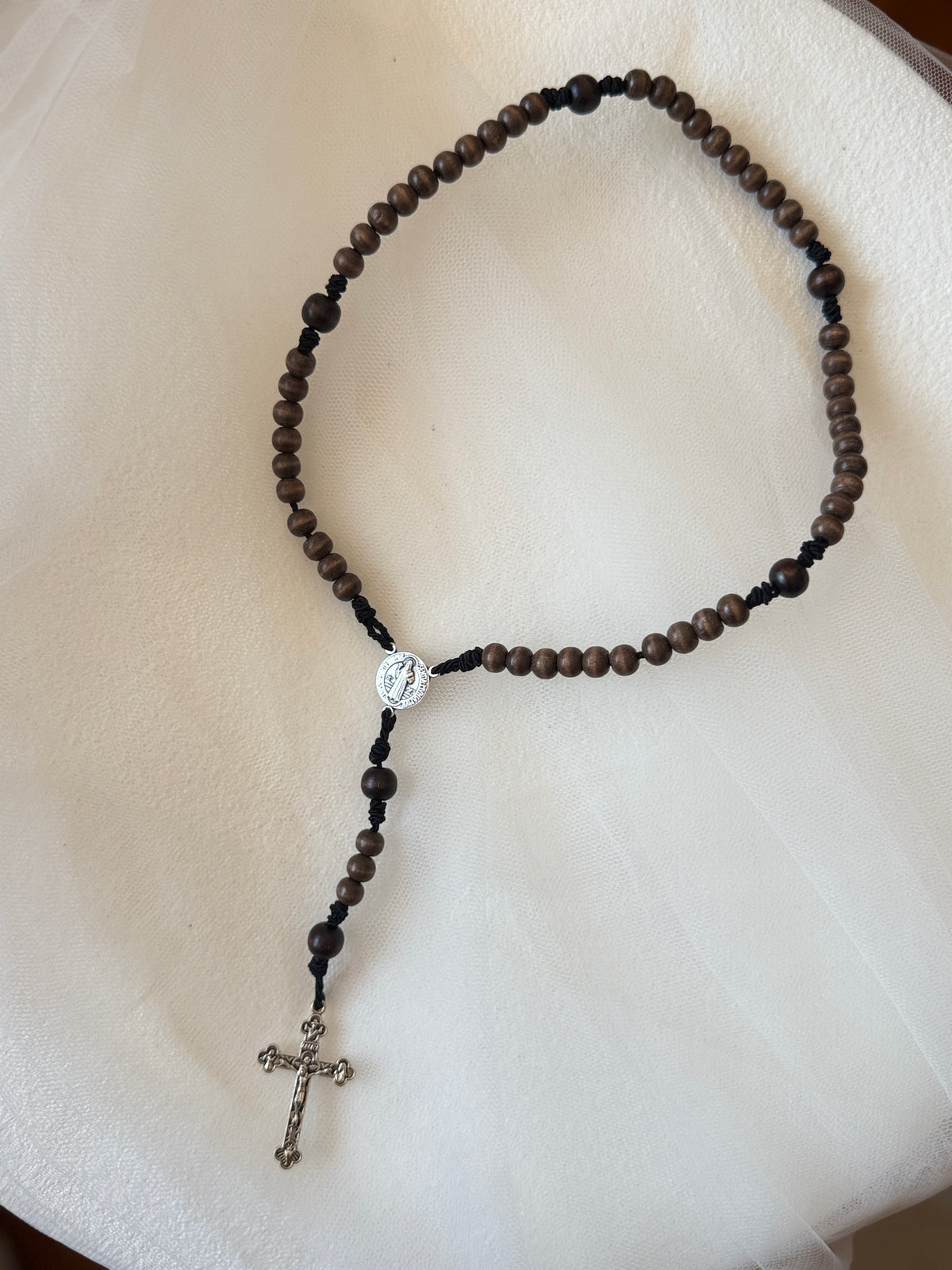 A dark brown wooden bead rosary necklace with a silver-tone centerpiece medallion and a dangling silver cross, displayed on a soft white fabric background.
