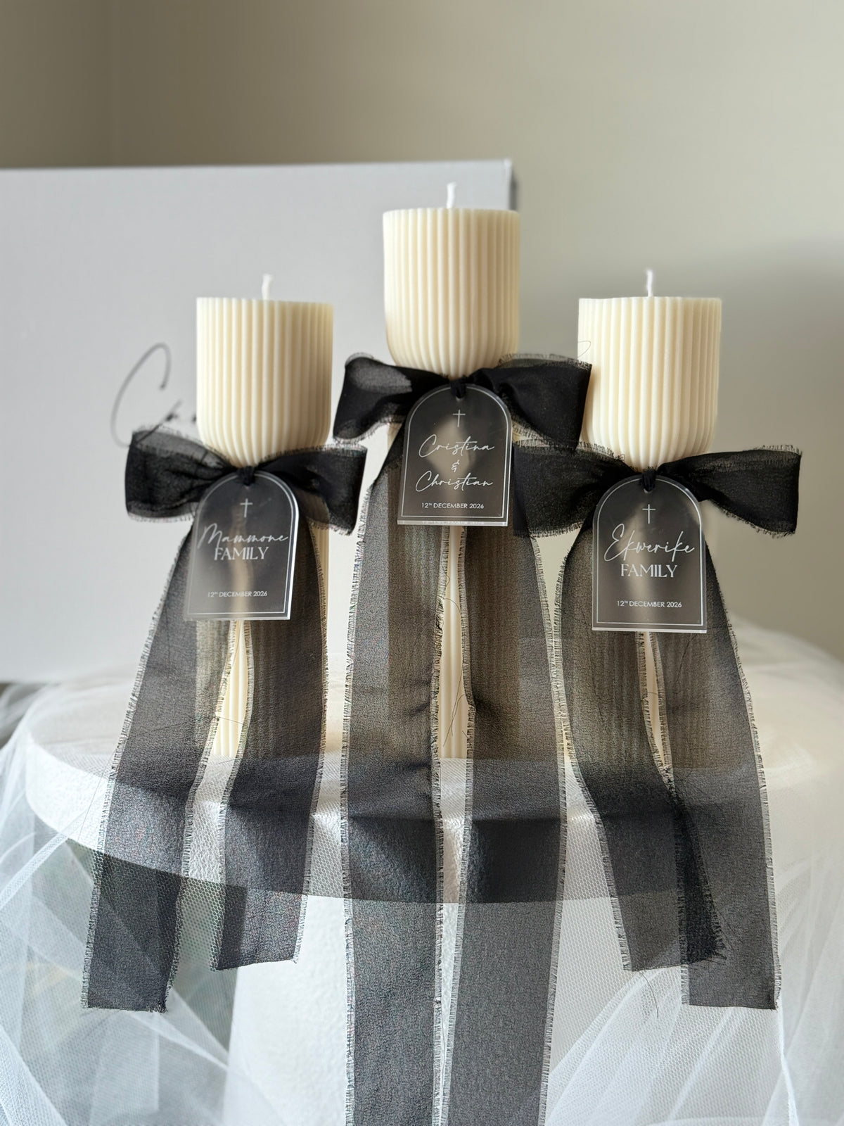 Three cream-colored ribbed pillar candles on sticks, each tied with a black sheer ribbon bow and a small hanging tag, displayed on a white surface with a soft neutral background.
