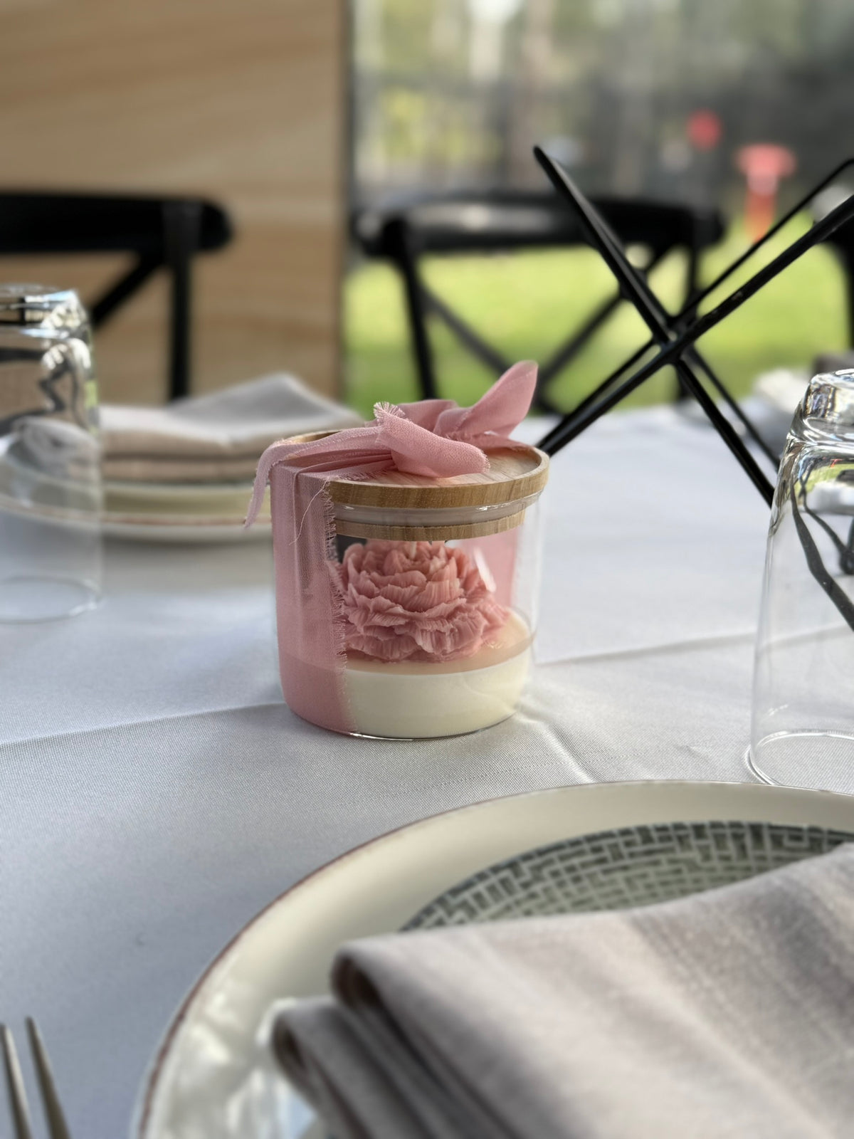 A small glass jar candle or decorative container with a pink flower-shaped wax piece inside, tied with a pink ribbon, set on a white tablecloth in a styled dining setting.