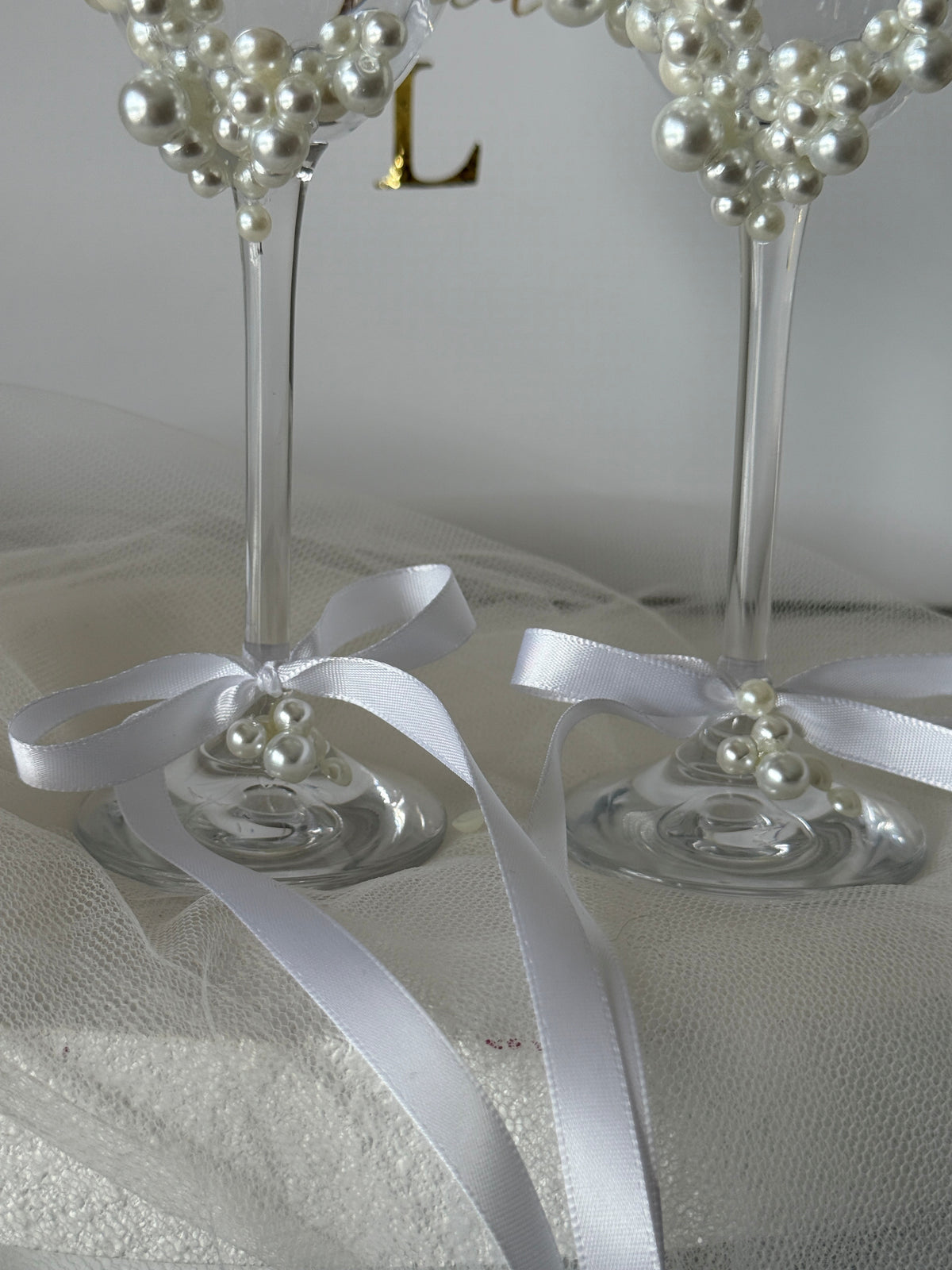 Two clear glass highball glasses decorated with clusters of white faux pearls at the top and tied with white satin ribbons at the base, displayed on a white fabric surface.