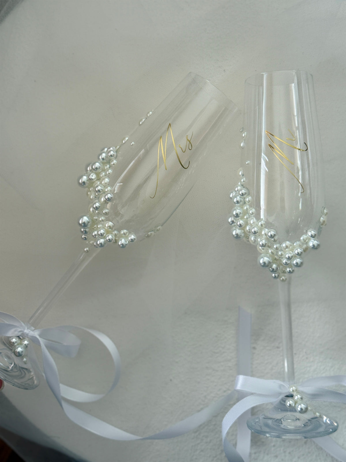 Two clear champagne flutes decorated with white pearl bead clusters, gold script lettering, and white satin ribbons, shown on a light neutral background.