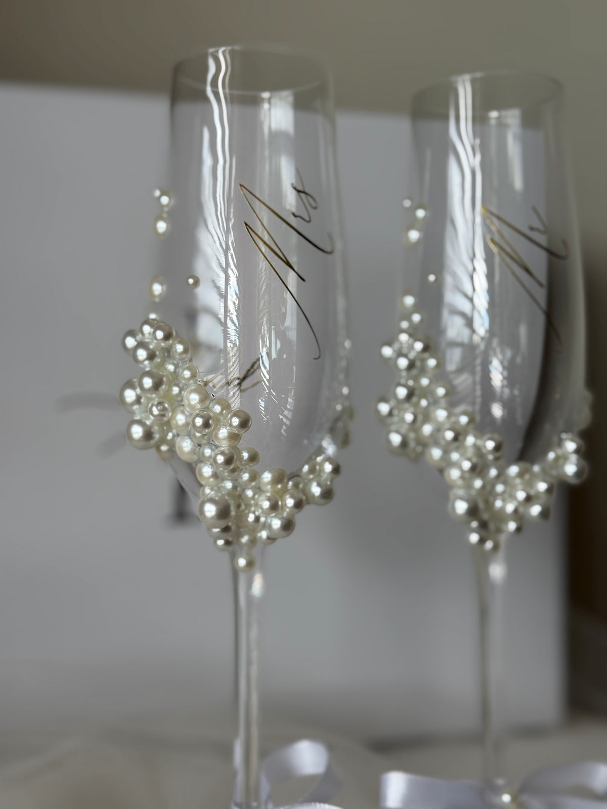 Two clear champagne flutes decorated with white faux pearls and gold script lettering, shown against a soft neutral background.