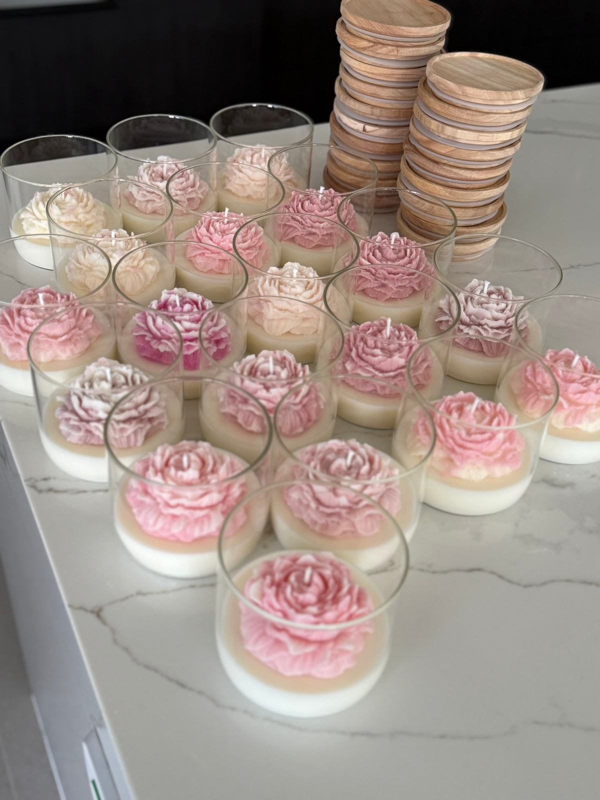 Multiple clear cylindrical glass containers filled with layered white and pink rose-shaped wax melts or soaps, arranged on a white marble countertop with stacked wooden lids in the background.