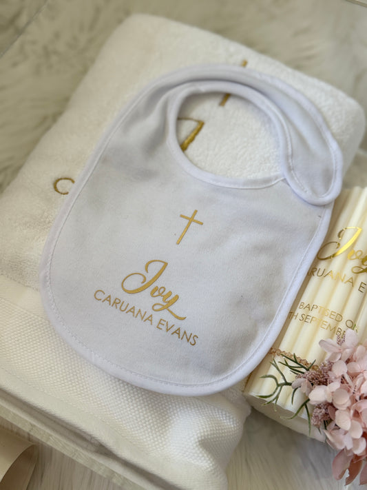 A white personalized baby bib with gold embroidery featuring a cross, the word 'Joy', and the name 'CARUANA EVANS', styled on a white towel next to a candle and pink flowers.