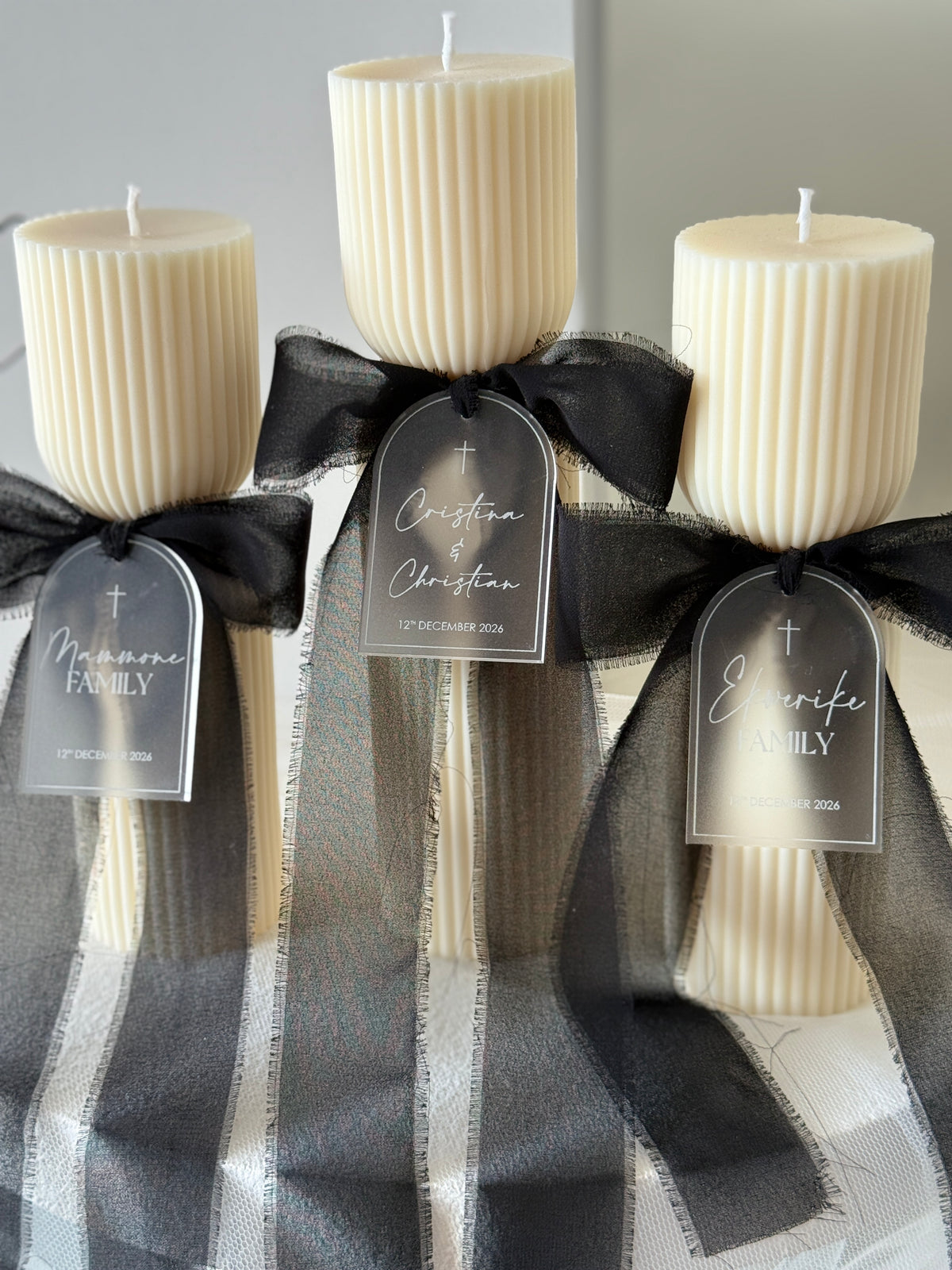 Three cream-colored ribbed pillar candles tied with black sheer ribbon and personalized tags, wrapped in a black and white plaid fabric at the bottom.