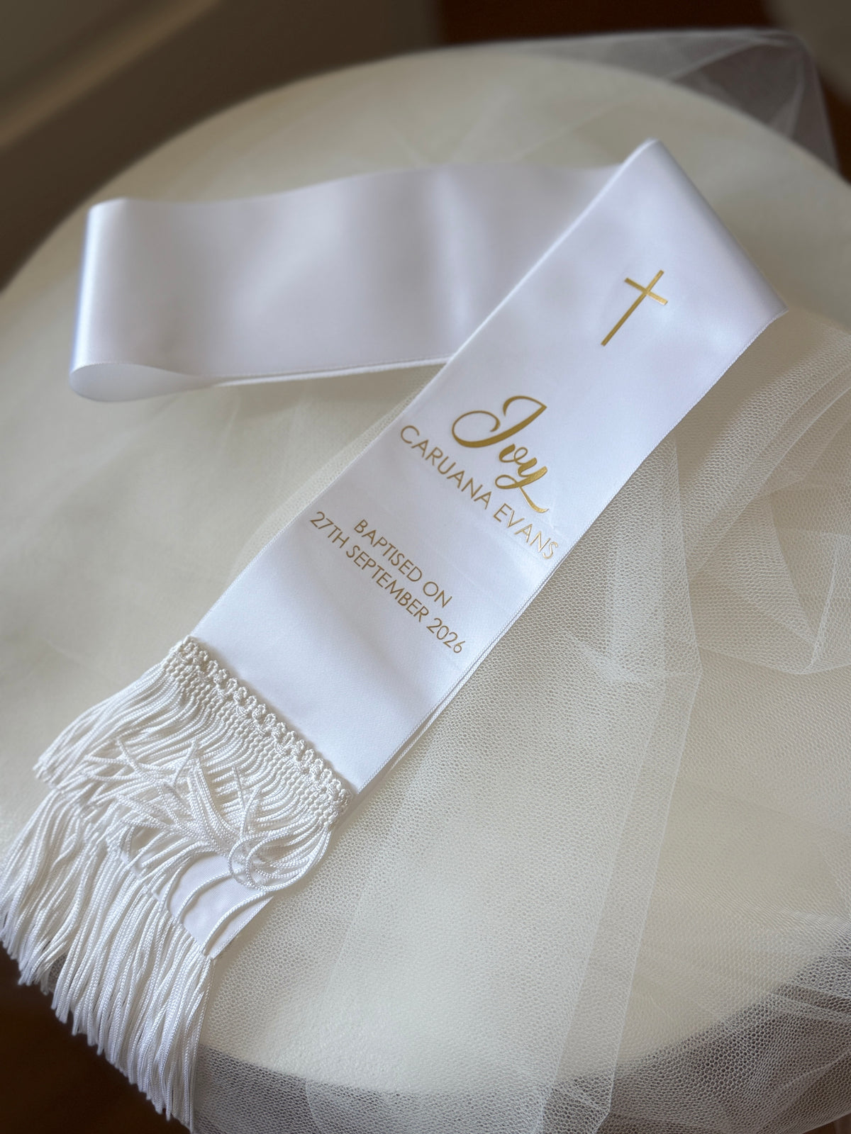 A white satin baptismal or confirmation sash with gold lettering and a gold cross, laid on a round white table covered with sheer white tulle.